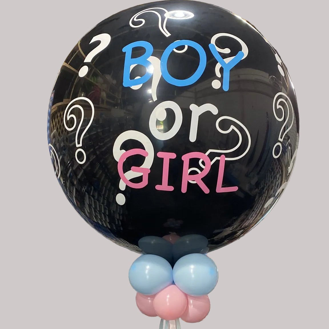 A large black party balloon printed with the words 'Boy or Girl' in blue, white, and pink lettering, with question marks and swirls, standing in a retail store aisle with shelves of packaged items in the background.