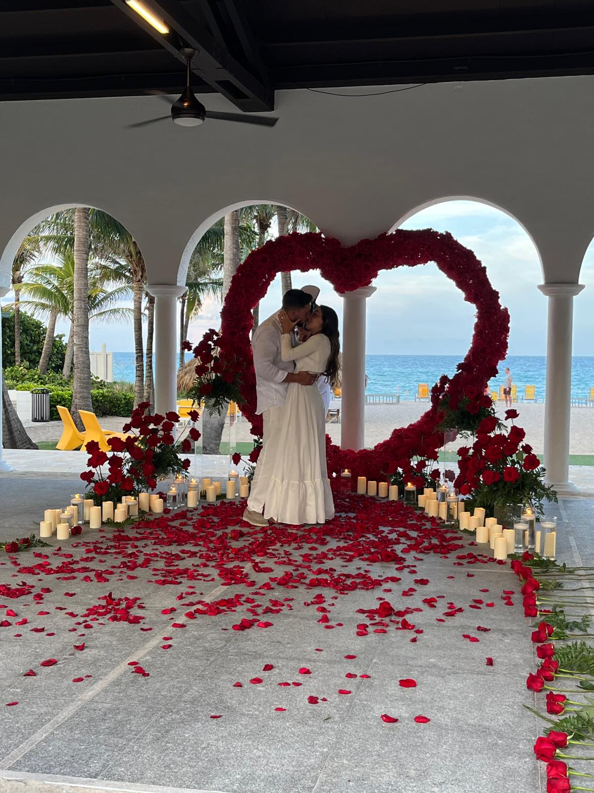 Couple embracing under a heart-shaped arch decorated with flowers and candles, with a beach and palm trees in the background.