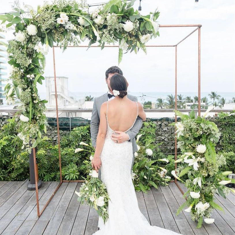 Couple standing under a floral arch on a wooden deck with greenery around.