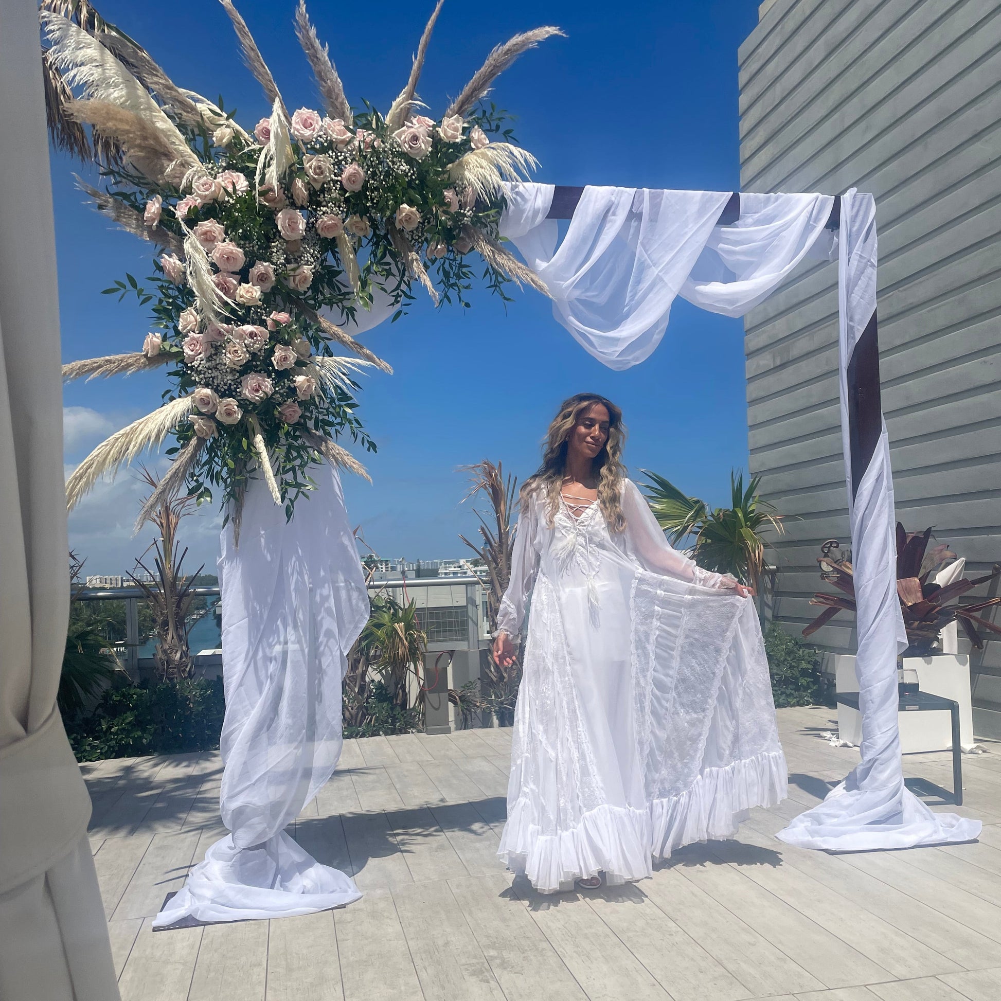 Woman in a white dress standing under a floral arch on a wooden deck with a clear blue sky.