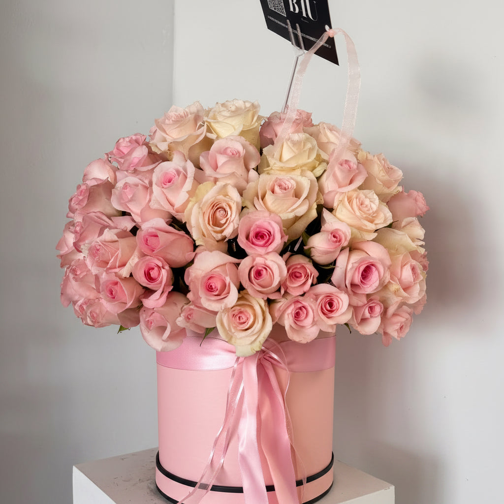 A large bouquet of pink and cream roses arranged in a round pink box with a pink ribbon, displayed on a white pedestal against a light gray wall.